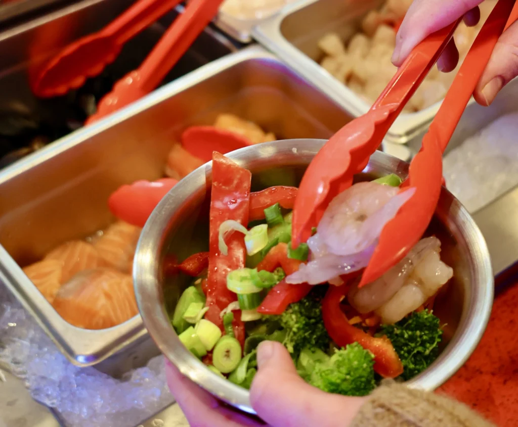 closeup of hand holding bowl of ingredients and tongs adding shrimp to bowl at buffet