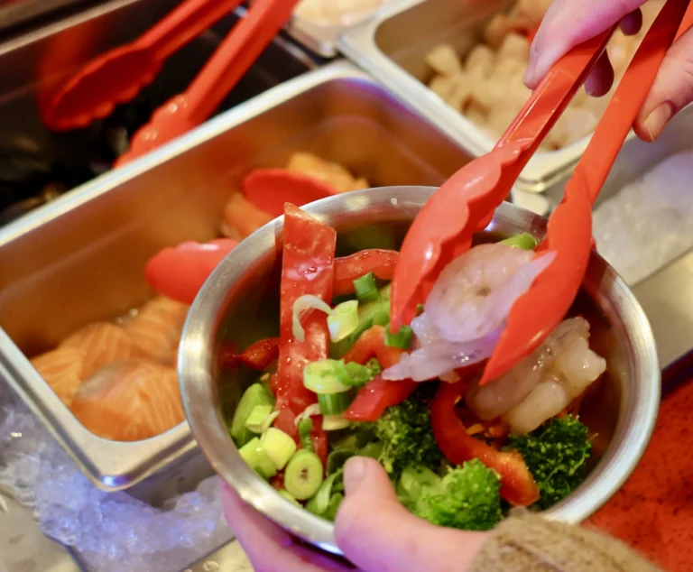 closeup of hand holding bowl of ingredients and tongs adding shrimp to bowl at buffet